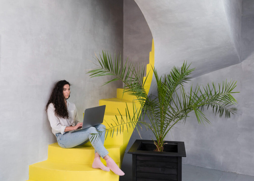 mujer sentada en escalera amarilla trabajando en portatil en espacio moderno minimalista con planta decorativa